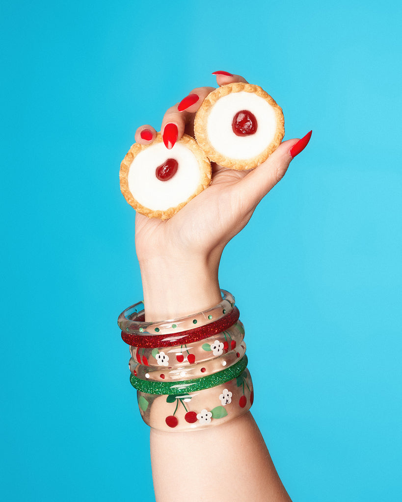 Splendette vintage inspired 1950s rockabilly style Cherries Clear Bangles with red and green glitters modelled by a hand holding Bakewell tarts